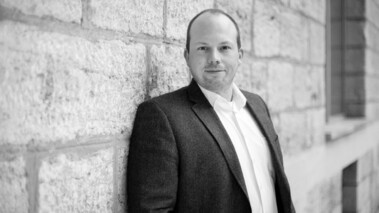 Porträt von Sebastian Klembt in blauem Sakko vor einer Sandsteinwand, leicht lächelnd.  Dateiname: reinraumlabor-video-screenshot-20210226.jpg